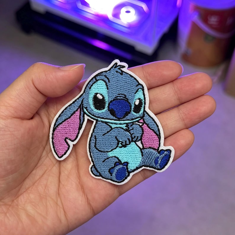 May include: An embroidered patch featuring the character Stitch in shades of blue and light blue with pink inner ears, a dark blue nose, and black eyes. The patch has a white border and depicts the character in a seated position with arms folded.