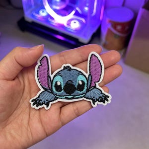 以下が含まれることがあります： 青い毛並み、大きな紫色の耳の内側、黒い目を持つキャラクター「スティッチ」の刺繍ワッペン。デザインは白い糸で縁取られており、キャラクターが腕を前に伸ばして伏せている様子が描かれています。