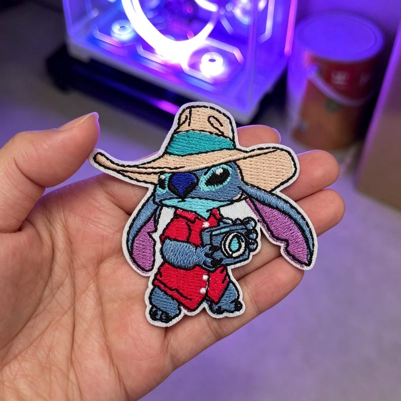 May include: An embroidered patch featuring a blue alien character wearing a tan hat with a teal band and a red shirt. The character is holding a small blue camera with a white lens detail. The patch has a white border and purple inner ears.