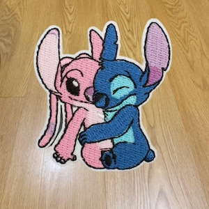 May include: An embroidered patch featuring two cartoon characters in a close embrace. One character is light pink with large ears, while the other is blue with darker blue accents and large ears. The design is outlined in white thread.