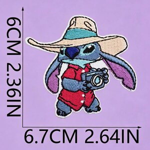 May include: An embroidered patch featuring a blue alien character wearing a tan sun hat and a red shirt while holding a camera. The patch measures 6 cm by 6.7 cm, or 2.36 inches by 2.64 inches, as indicated by the black measurement lines.