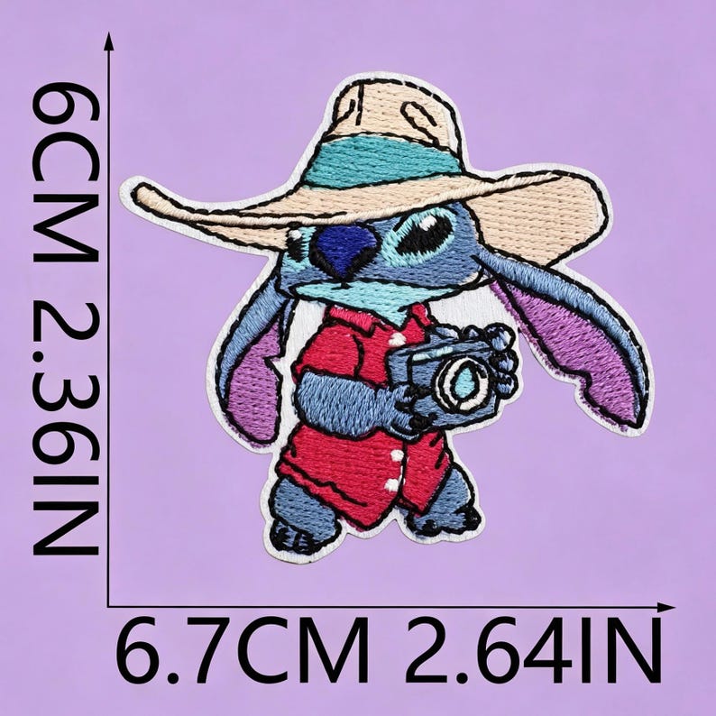 May include: An embroidered patch featuring a blue alien character wearing a tan sun hat and a red shirt while holding a camera. The patch measures 6 cm by 6.7 cm, or 2.36 inches by 2.64 inches, as indicated by the black measurement lines.