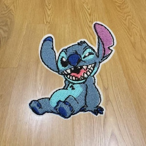 Puede incluir: Un parche bordado que muestra al personaje Stitch en posición sentada. El diseño utiliza tonos de azul para el cuerpo, una nariz azul oscuro y el interior de las orejas rosa. El personaje tiene una amplia sonrisa con dientes hecha en hilo.