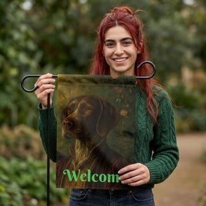 Op de afbeelding: Een tuinvaandel met een portret van een bruine hond en het woord "Welcome" in het groen. De vlag wordt vastgehouden door een zwarte metalen standaard, ideaal voor buitendecoratie.