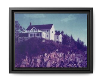 Puede incluir: Impresión en lienzo enmarcada que presenta una fotografía de estilo vintage de una casa en un acantilado. La casa blanca tiene un tejado oscuro, sobre un cielo azul. El primer plano muestra follaje púrpura y terreno rocoso. El marco es negro.