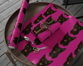 Könnte beinhalten: Rolle rosa Geschenkpapier und ein verpacktes Geschenk mit brauner Schleife, beide mit einem sich wiederholenden Muster aus schwarzen Katzenköpfen mit herausgestreckter Zunge. Schere und Klebeband liegen ebenfalls auf dem Tisch.