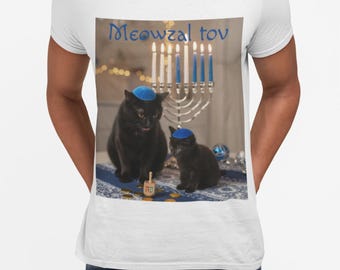 May include: White t-shirt featuring a graphic of two black cats wearing blue yarmulkes, with a menorah and dreidel. The text "Meowzal Tov" is written above the image in blue. The cats are on a blue and white patterned surface.
