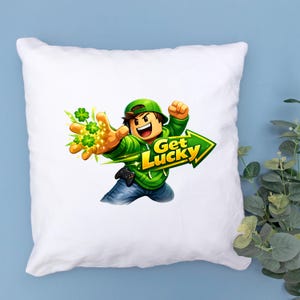 Op de afbeelding: Wit vierkant kussen met een stripfiguur in een groene hoodie en hoed, die een hand met klavers uitsteekt. Een groene pijl met de woorden "Get Lucky" wijst naar rechts. Het personage draagt een blauwe spijkerbroek.