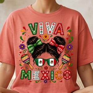 Pode incluir: Uma t-shirt cor de coral com uma impress&atilde;o gr&aacute;fica de cabelo escuro em dois coques com la&ccedil;os verdes e vermelhos, &oacute;culos de arma&ccedil;&atilde;o verde com a bandeira do M&eacute;xico e duas maracas. O texto VIVA MEXICO est&aacute; impresso em letras decorativas.