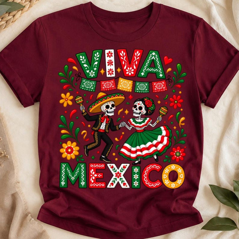 Pode incluir: Uma t-shirt de manga curta cor de vinho com um gr&aacute;fico de dois esqueletos a dan&ccedil;ar. O texto VIVA MEXICO est&aacute; impresso em letras grandes e decorativas. O design inclui maracas, flores e bandeirolas coloridas em vermelho, verde e amarelo.