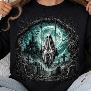 Puede incluir: Una sudadera negra con un estampado gráfico de un fantasma blanco sentado en un columpio. La escena incluye una gran luna llena, un castillo gótico oscuro, un cementerio con lápidas y ramas de árboles retorcidas que enmarcan la ilustración.
