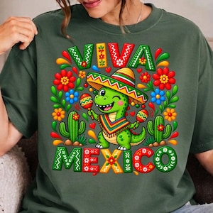 Pode incluir: Uma t-shirt verde-oliva escura com um gr&aacute;fico de um dinossauro verde usando um sombrero e um poncho coloridos, segurando maracas. O design inclui cactos, flores e o texto VIVA MEXICO em letras de bloco grandes e multicoloridas.