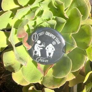 May include: A round, black button with white text and graphics, reading "Derby: Cheaper Than Therapy." The button features stick figures and is set against a backdrop of a green succulent plant.