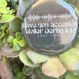 May include: A dark gray, round object with the text "Have you accepted Roller Derby into your life?" in white. The object is surrounded by green succulent plants. A white sound wave graphic is above the text.