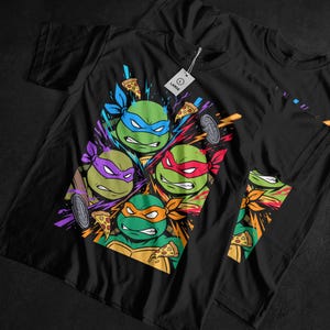 May include: Black t-shirts featuring a colorful graphic of the Teenage Mutant Ninja Turtles. Each turtle is depicted with their signature mask color, surrounded by pizza slices and comic book style effects. The shirts have a "LARGE" tag.