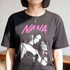 May include: Black t-shirt featuring a graphic of a woman with short hair, a microphone, and the word "NANA" in pink. The illustration is in a manga style. The woman is wearing a choker and a corset-style top.