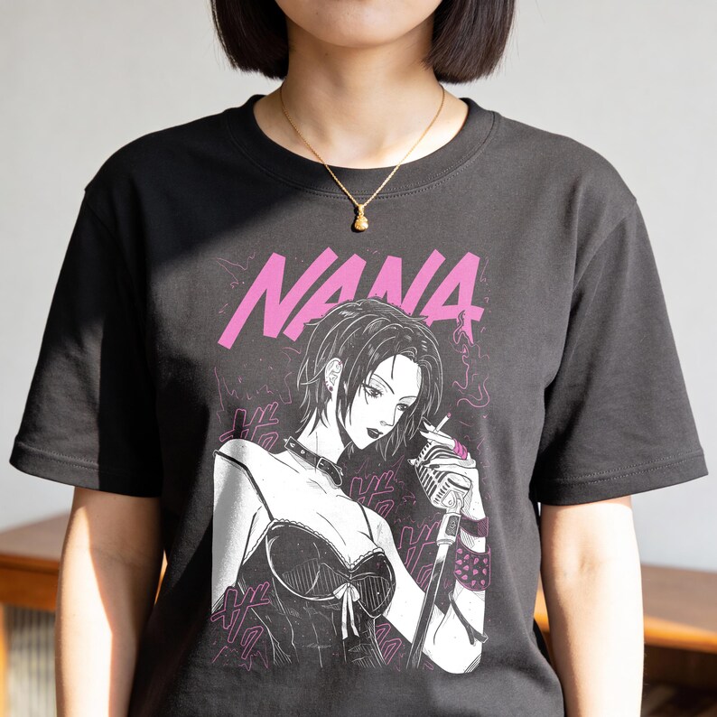 May include: Black t-shirt featuring a graphic of a woman with short hair, a microphone, and the word "NANA" in pink. The illustration is in a manga style. The woman is wearing a choker and a corset-style top.