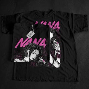 May include: Black t-shirts featuring a manga-style illustration of a woman with the word "NANA" in pink. One shirt is folded, with a tag that reads "LARGE". The design includes a microphone and smoking imagery.