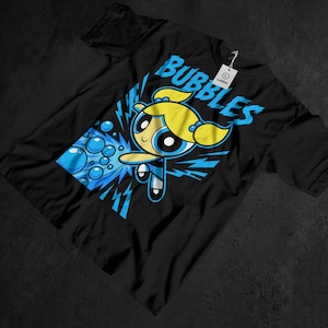 Op de afbeelding: Zwart t-shirt met een cartoonfiguur met geel haar en een blauwe outfit. Het woord "BUBBLES" staat in het blauw boven het personage gedrukt. Het ontwerp bevat blauwe bellen en bliksemschichten.