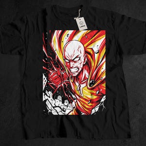 May include: Black t-shirt featuring a vibrant graphic of a bald, muscular character with an intense expression. The artwork uses red, yellow, and white colors, with a dynamic, action-oriented design. A tag with the word "LARGE" is attached.