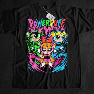 Op de afbeelding: Zwart t-shirt met een kleurrijke afbeelding van de Powerpuff Girls en het woord "POWERPUFF". Het ontwerp toont Bubbles, Blossom en Buttercup. Het shirt heeft een "LARGE" label.