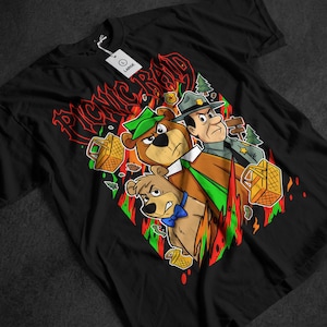 Pode incluir: Camiseta preta com um desenho animado de dois ursos e um guarda florestal, com o texto "PICNIC RAID" em vermelho. Os ursos estão segurando cestas de piquenique, e o guarda florestal está usando um chapéu. O design tem uma borda em forma de diamante com detalhes verdes e vermelhos.
