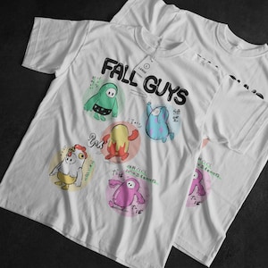 Puede incluir: Camisetas blancas con el texto "FALL GUYS" e ilustraciones de personajes de dibujos animados coloridos. Los diseños incluyen varios personajes en diferentes colores, con texto japonés y una etiqueta "LARGE".