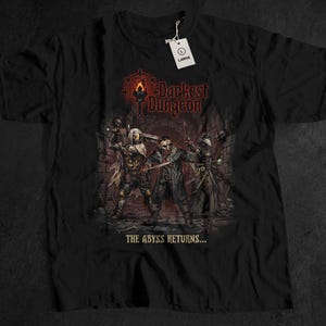 Peut inclure: T-shirt noir avec un graphisme détaillé du jeu Darkest Dungeon. Le design présente trois personnages armés, le logo du jeu et le texte "THE ABYSS RETURNS...". Une étiquette blanche avec "LARGE" est attachée.