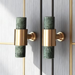 Op de afbeelding: Close-up van meubelbeslag met cilindrische groene marmeren handgrepen met goudkleurige metalen accenten. De handgrepen zijn gemonteerd op een lichtgrijze houten kastdeur met gouden afwerking.