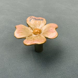 May include: A drawer knob shaped like a four-petaled flower with a metallic gold finish and soft pink enamel shading on the petals. The centre of the flower has a textured gold appearance. The text Thomas is printed on one petal with a dolphin icon.