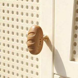 Puede incluir: Un pomo de armario de madera con forma ovalada y tres secciones talladas. El pomo está sujeto a una puerta de armario blanca con una base metálica de color latón. La puerta del armario tiene un panel perforado de color beige claro.