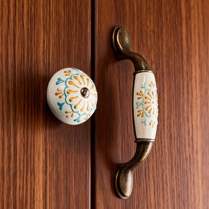 May include: A white ceramic cabinet knob and handle set with a floral design in gold and teal. The hardware has a bronze finish and is mounted on a wooden cabinet door.