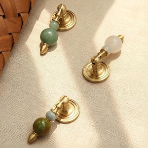May include: Three decorative cabinet pulls with brass hardware and gemstone accents. The pulls feature a round base, a curved arm, and a gemstone sphere. The gemstones are in shades of green and white. The hardware has a gold finish.