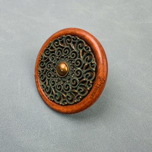 May include: A circular cabinet knob featuring a smooth, polished brown wooden outer ring and a central bronze-toned metal inlay with an intricate, swirling filigree pattern. A small, smooth bronze sphere sits at the center, and the name Thomas is engraved on the wood.