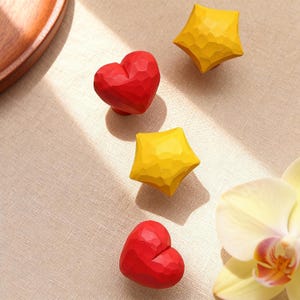 May include: Red heart-shaped and yellow star-shaped decorative knobs. The knobs have a faceted design and are arranged on a neutral-colored surface. The heart knobs are red, and the star knobs are yellow.
