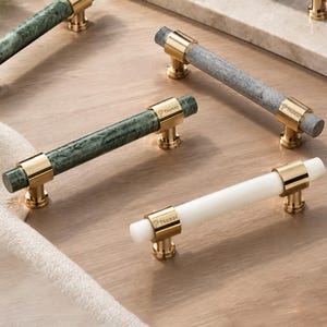 以下が含まれることがあります： 緑、グレー、白の石材を使用した円筒形のバーを備えたキャビネットプルコレクション。それぞれが磨かれたゴールドトーンの金属製マウントでアクセントが付けられています。ゴールドのマウントにはブランド名Thomasが刻印されています。