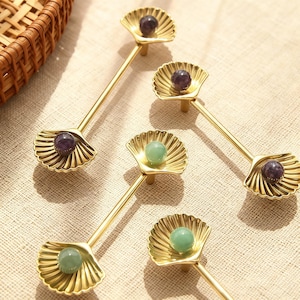 May include: Four decorative cabinet pulls with a seashell design. The pulls are gold-colored with a small, round gemstone in the center of each shell. Two pulls have purple gemstones, and two have green gemstones.