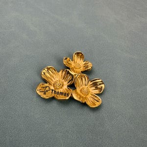 Puede incluir: Un broche de metal dorado que presenta tres flores conectadas con centros texturizados. Los pétalos tienen un acabado liso y reflectante. El nombre Thomas está grabado en negro en uno de los pétalos, junto a un pequeño icono de un delfín.