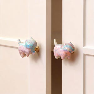 May include: Two cat-shaped cabinet knobs. The knobs are pink, blue, and white with a marbled design. The knobs are attached to a white door with gold-colored hardware. The cat knobs are a decorative home decor item.