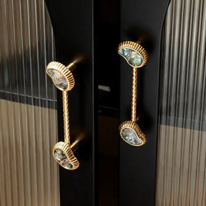 Puede incluir: Primer plano de una puerta de armario negra con dos tiradores dorados. Cada tirador presenta un vástago retorcido y un extremo decorativo con una incrustación de concha de abulón. La puerta del armario tiene un panel de vidrio acanalado.