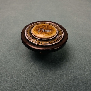 May include: A circular cabinet knob featuring a dark brown wooden outer ring, a metallic band with a Greek key pattern, and a central amber-colored stone inlay. The word Thomas is printed in white text across the center of the stone.