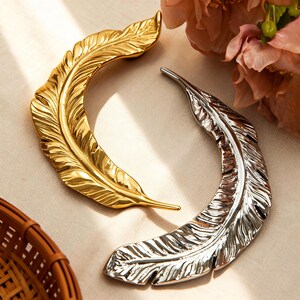 Può includere: Due oggetti decorativi a forma di piuma, uno dorato e l'altro argentato. Entrambi presentano dettagliate texture di piume e una forma curva. Sono esposti su una superficie neutra, con un cestino intrecciato e fiori sullo sfondo.