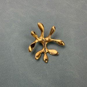 May include: A polished gold-toned metal object with an irregular, organic, multi-lobed shape. The surface features a small engraved dolphin icon and the name Thomas printed in black lettering, positioned centrally on the smooth, reflective metal.