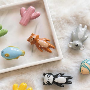 Könnte beinhalten: Eine Sammlung farbenfroher Keramik-Tierfiguren, darunter ein rosa Kaktus, ein Fuchs, ein Koala, ein Panda und ein blauer Fisch. Die Figuren sind auf einem weißen Tablett und einer flauschigen weißen Oberfläche angeordnet.