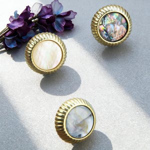 May include: Three decorative cabinet knobs with gold-colored, ridged edges. The center of each knob features a pearlescent, iridescent, or white inlay. A sprig of purple flowers is in the background.