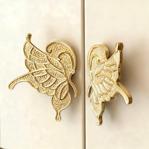 May include: Two gold butterfly-shaped cabinet pulls. The metal pulls feature detailed wing designs. The cabinet pulls are mounted on a light-coloured cabinet door.