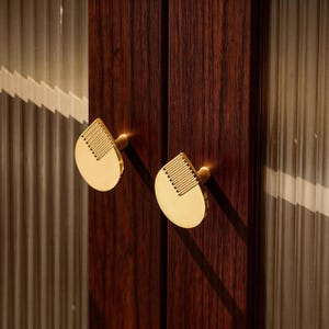 May include: Close-up of a pair of gold-colored cabinet door pulls. The pulls have a teardrop shape with a series of parallel lines on one side. The cabinet doors are dark wood with a vertical grain. The background has a textured, translucent panel.