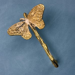 May include: A gold-toned metal butterfly-shaped object with a long, slender, textured handle. The word Thomas is engraved on the butterfly wing and again on the handle. The design features intricate wing patterns and a polished metallic finish.