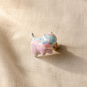 Könnte beinhalten: Ein katzenförmiger Möbelknopf mit einem Marmormuster aus pastellfarbenen rosa, blauen und weißen Wirbeln. Der Knopf hat eine glänzende Oberfläche und einen goldfarbenen Metallsockel. Das Wort Thomas ist in kleinen weißen Buchstaben seitlich aufgedruckt.
