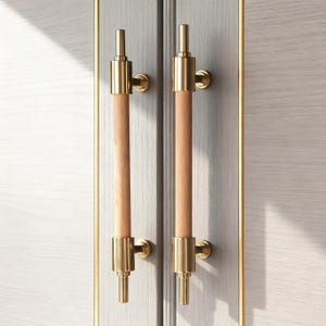 May include: Two cabinet door handles with a light wood grain centre and gold-coloured metal accents. The handles are cylindrical with a vertical design, mounted on a light grey cabinet door with a gold trim.